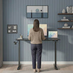 Femme debout travaillant de dos sur un bureau assis-debout électrique noir réglé en hauteur, avec un grand écran et un ordinateur portable, dans un bureau à domicile moderne au mur bleu