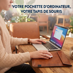 Une femme travaille sur son ordinateur portable dans un café. Sa pochette d'ordinateur en cuir marron est ouverte sur la table en bois et sert de tapis de souris pratique. En haut de l'image, le texte dit : VOTRE POCHETTE D'ORDINATEUR, VOTRE TAPIS DE SOURIS.