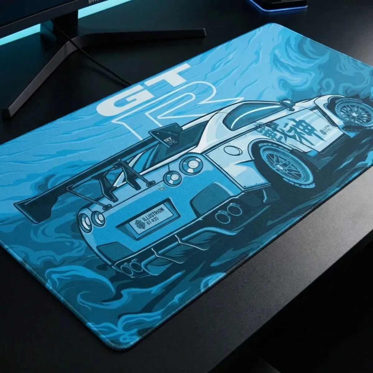 Gros plan sous un angle oblique du Tapis de souris Apex Skyline GT-R bleu clair sur un bureau noir, avec moniteur et clavier. Montre la texture micro-texturée et l'illustration GTR-R35. Format XXL.