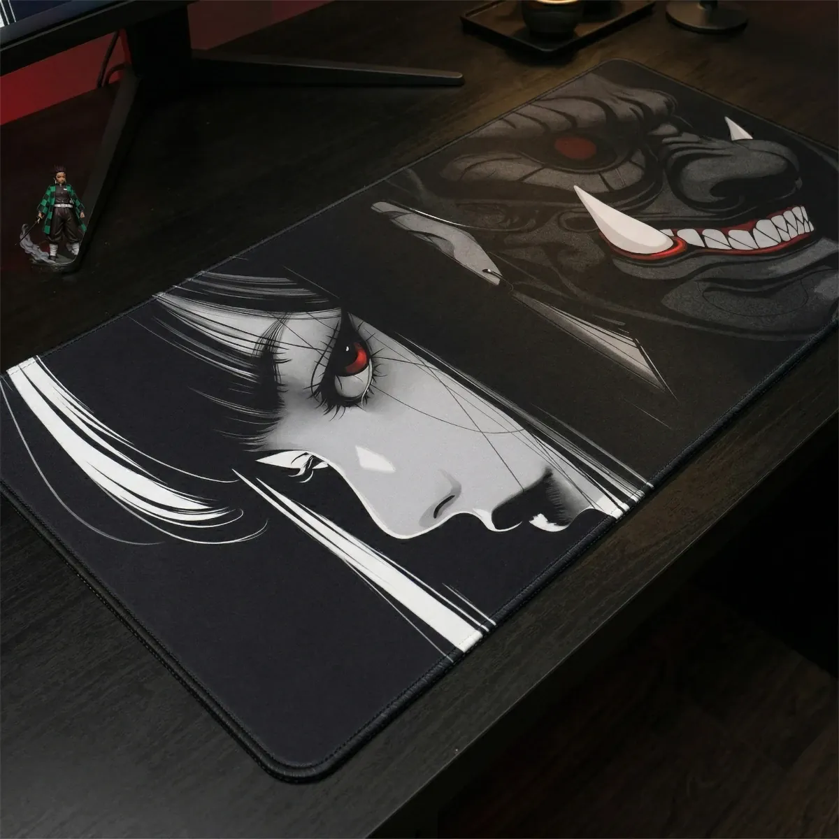 Gros plan du tapis de souris Oni Phantom XXL sur un bureau en bois noir, montrant son design de masque Oni et portrait de femme, avec une figurine Demon Slayer et des accessoires de bureau à côté.